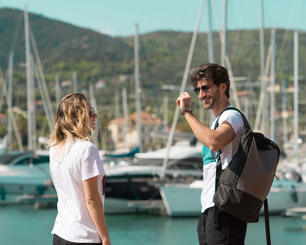 Two travelers happily converse near the boats. Man wears a gray DRiiBE backpack